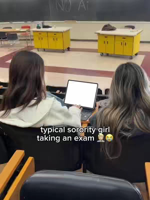 The image shows two young women from behind, seated in a classroom setting with a laptop between them. The foreground shows a chair and desk details. The background includes yellow cabinets and a chalkboard, suggesting a college classroom environment.