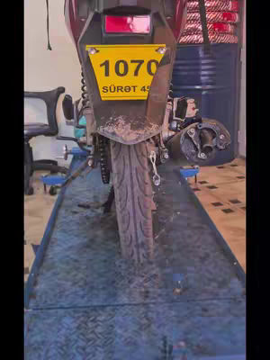 The image shows the rear of a motorcycle, positioned on a blue metal platform with a diamond pattern. The motorcycle is primarily dark red, with a visible tire and license plate featuring the number '1070' and the text 'SÜRƏT 45' in yellow. The background includes what appears to be an industrial setting, with elements like a large metal drum. The composition is a straight-on shot, emphasizing the motorcycle's details and the functional setting.
