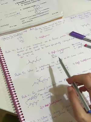 A top-down shot shows a person's hand holding a pen, writing on a notebook filled with handwritten notes and diagrams. A textbook is visible in the background.