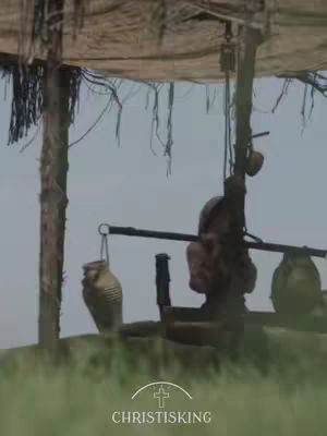 The image shows a desert scene with a well and shaded structure. A long wooden beam is used for the well, with a large jar on either side for water. The foreground features green grass, and the background shows a clear sky.