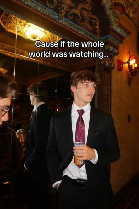 A man in a black suit with a burgundy tie and a drink in his hand stands in a room with ornate decor and a large mirror. Text overlays the image at the top.