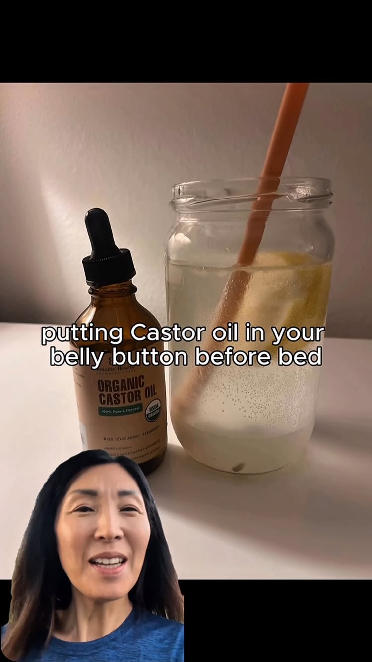 The cover image shows a woman smiling while she is standing in front of a table with a glass jar on the table. The jar has a straw inside and is approximately half full with a clear liquid, and there is a lemon slice inside. Next to the jar is a bottle of brown liquid, with a black top, with text labels, and the main text is 'ORGANIC CASTOR OIL'. A text overlay on the image states 'putting Castor oil in your belly button before bed'.