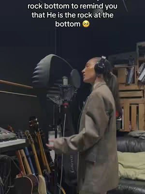 A woman stands in a recording studio, singing into a microphone. She wears a blazer, and headphones. Behind her is a microphone and soundproofing. In the background are musical instruments