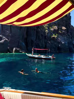 The photo captures a serene scene of a boat trip on a sunny day. A striped yellow and red awning casts shade over the top of the frame. Beneath, vibrant turquoise water stretches out, with several figures swimming. A small boat with people on board floats in the middle distance, and rocky cliffs form the backdrop.
