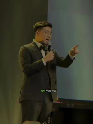 A man in a suit and bow tie stands on a stage with a microphone in hand, pointing to his right. The background is a mix of light and dark colors.