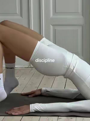 A woman in white workout clothes is doing a bridge exercise on a gray yoga mat. Her legs are raised, and her arms are extended on the mat for support. The word "discipline" is overlaid on the image in white text.