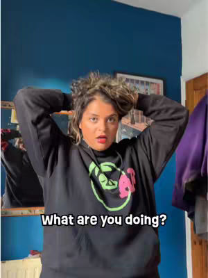 A woman with curly brown hair and a black hoodie with a logo looks directly at the camera with a surprised expression. The background includes a blue wall, a mirror, and a door. The overlay text at the bottom reads, 'What are you doing?'