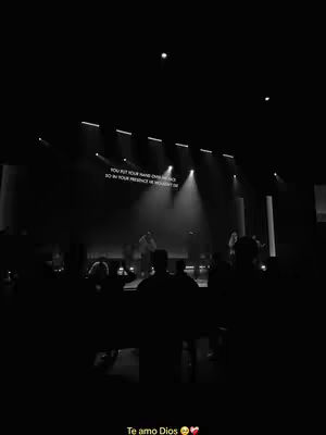 A low-angle, black and white photograph captures a dimly lit church interior during a worship service. Silhouetted figures of the congregation fill the foreground, facing towards a stage. On the stage, figures are partially visible, likely the worship team, illuminated by spotlights. Above them, a screen displays white text against a dark background. The overall atmosphere is serene and reverent.