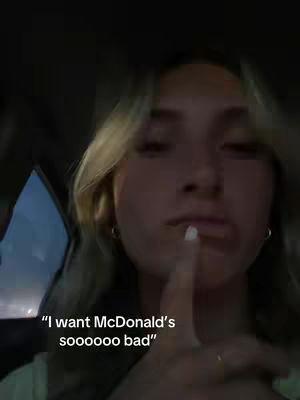 A young woman in a car is looking towards the camera, pointing her index finger up to her lips. The background is dark, with some visible car interior elements. Text overlaid on the image reads: "I want McDonald's soooooo bad".