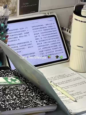 A well-lit, close-up shot of a student's desk displaying open books, a tablet, and study materials. A notebook and some papers are in the foreground, partially obscuring a laptop. A pen rests near the papers. The tablet screen displays what appears to be a document, The background includes a water bottle and a decorative item.