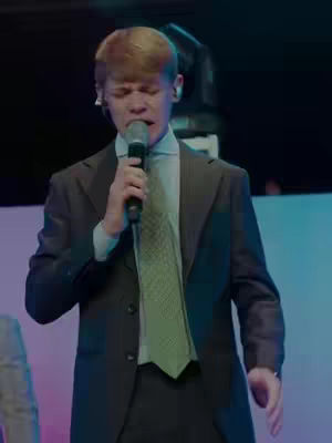 A man in a suit singing into a microphone. The background is blurry with colored lights. The man is wearing a green tie and has an earpiece.