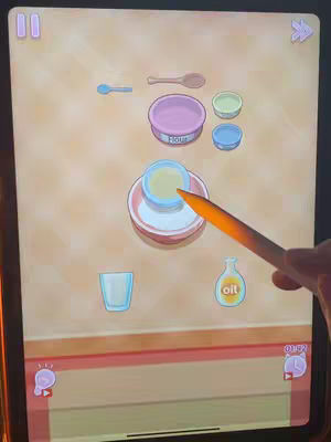 The cover image depicts a cooking game interface on a tablet, showing various cooking tools like bowls, spoons, and bottles of oil laid on a light-brown background. A hand holding a stylus is interacting with the screen, highlighting a step in the cooking process using an orange stylus.