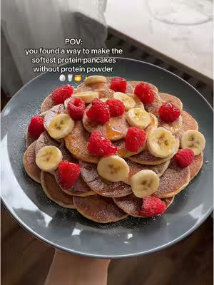 A close-up shot showcases a plate filled with meticulously arranged pancakes, adorned with fresh raspberries and banana slices. The pancakes, golden brown and round, are layered in a slight spiral, interspersed with bright red raspberries and creamy yellow banana slices. The plate holding the pancakes is dark gray.