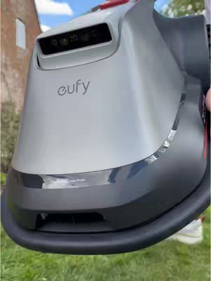 The cover image shows a close-up of a robotic lawnmower. The majority of the view is of the mower's top, which is a smooth, metallic gray. The front portion is a darker, almost black color, with a line of text in a reflective panel. The 'eufy' logo is visible in the center. The background is blurred, but we can see grass and a building.