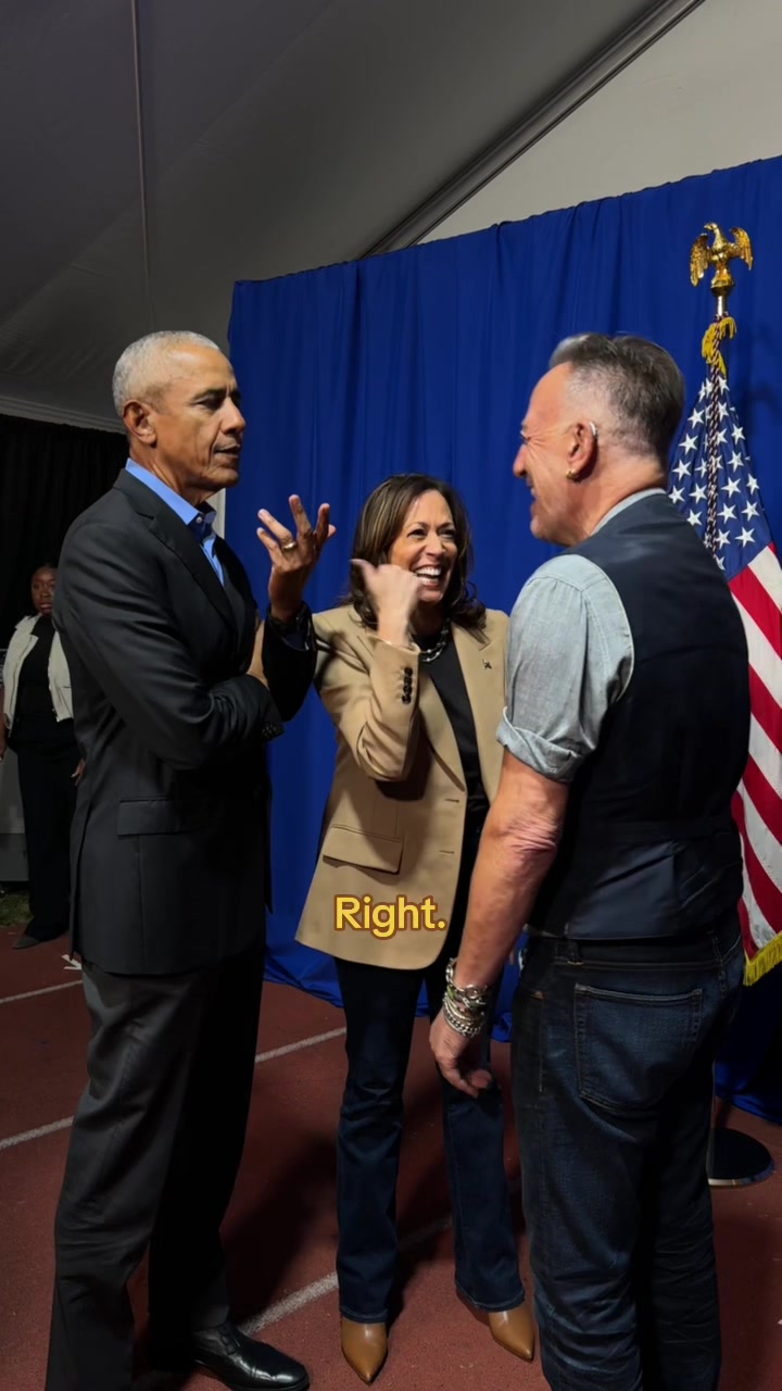 A colorful image captures Barack Obama, Kamala Harris, and Bruce Springsteen in a candid moment. Obama, in a sleek black suit, is seen gesturing with one hand while speaking to Harris. Harris, centrally positioned with a business jacket, is laughing. Springsteen, to the right, gazes at Harris with a smile. The background features a blue event backdrop, an American flag, creating a balanced composition. The image suggests a friendly interaction.