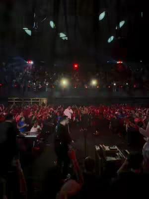An indoor, low-light shot of a live music event or religious gathering. The stage is in the foreground with a person in a black outfit, possibly a performer or speaker, near a microphone and other equipment. In the center is a person in a white outfit. The audience in the background is densely packed, with many people raising their hands. Overhead lighting fixtures are visible at the top. The colors are dominated by reds and blacks with bright accents.