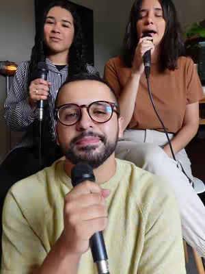 A group of three people singing into microphones. A man with glasses is in the foreground with two women in the background. The background behind them is blurred with a lamp.