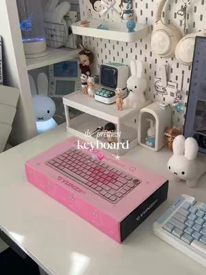 A well-lit photograph of a desk setup with a pink keyboard box as the main focus. The box sits on a white desk, and in the background, there is a shelf filled with various decorative items. The color scheme leans towards pastel pinks and whites, creating a cozy, aesthetic feel. A secondary keyboard and a white rabbit figurine are visible on the desk surface. The overall composition suggests an unboxing or desk setup video.