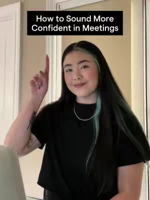 A woman with long dark hair is looking at the camera with a friendly expression over a background that has light-colored walls. She is pointing her index finger upwards. A banner with white text on a black background reads "How to Sound More Confident in Meetings".