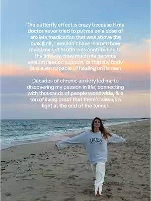 A woman stands on a sandy beach with her arms outstretched. The background is a blend of orange, and light blue as the sun sets. The woman is wearing a gray sweatshirt with the word "ARUBA" and white pants.