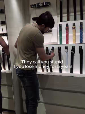 A man is standing in front of a display of belts, looking at them. The text overlay reads, 'They call you stupid if you lose money for 5 years'.