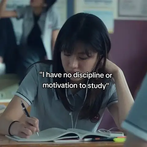 A young woman is seated at a desk, engrossed in writing in a notebook, with an earnest expression. The background shows a classroom setting with another student visible. A text overlay reads, " I have no discipline or motivation to study".