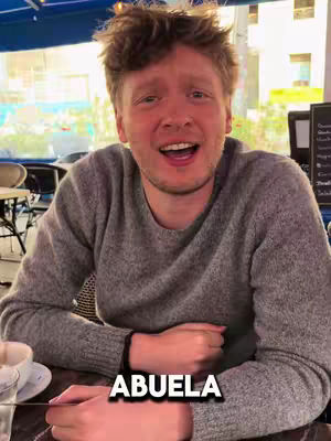 A man with reddish-blonde hair and a gray sweater sits at a table and smiles with his mouth open. Text overlay at the bottom says, 'ABUELA'. The background includes a blurred outdoor setting.