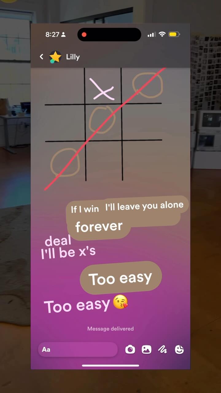 A phone screen displaying a text message conversation. A tic-tac-toe game is in progress, with the game grid visible. Text bubbles show the exchange between two people. The background shows the outside world.
