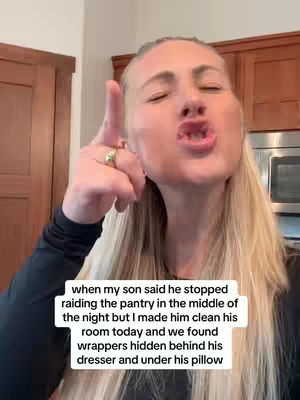 A woman with blonde hair is the main subject of this image. She points her index finger upwards prominently, with a slight frown and raised eyebrows. There's text overlaid in a white box at the bottom center detailing a story about a son and a pantry. The background is a home kitchen with wooden cabinets.