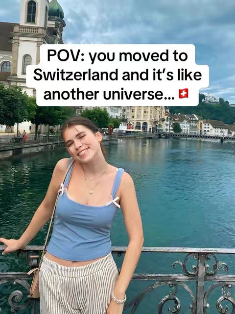 A young woman, wearing a blue top and striped trousers, stands on a metal railing, posing with a backdrop of a lake and buildings in Switzerland. Above her, a text overlay states, "POV: you moved to Switzerland and it's like another universe..." with the Swiss flag.