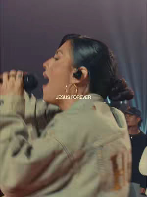 A woman in a beige jacket sings into a microphone, her mouth open and eyes closed. The text overlay 'JESUS FOREVER' is visible on the image. The background is blurred, with hints of stage lighting and other people.