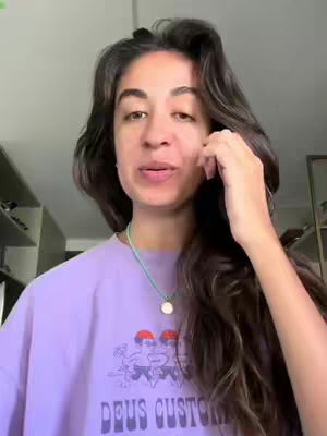 A medium shot shows a young woman with long, dark hair framing her face. She is wearing a purple t-shirt with a graphic. She is in a well-lit indoor space with shelves in the background.