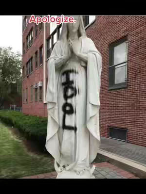 The image shows a white statue of a figure with hands clasped in prayer, possibly the Virgin Mary or a similar religious figure. The statue is vandalized with the word "FOOL" spray-painted in black across its front. The background consists of a reddish-brown brick building and a green lawn with trees.