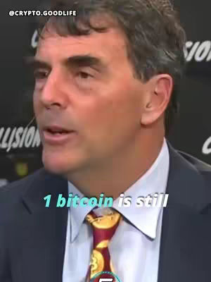 A man in a suit and tie is speaking, with the text overlayed near the bottom of the image "1 bitcoin is still". The background appears to be a conference or studio setting. The tie has a Bitcoin design in it.