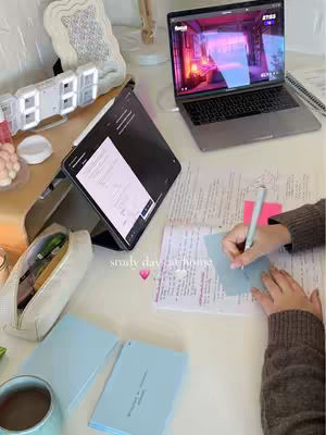 A top-down shot of a desk setup for studying. A laptop displays a colorful screen, while a tablet and notebooks are open. The scene is brightly lit, with a cup of coffee and a pen nearby. Hands are seen writing on a blue sticky note, with additional notes and study materials scattered around.
