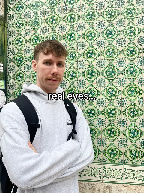 A man with light skin and short brown hair stands in front of a wall of green and white tile. The man wears a light grey hoodie and a black backpack. The text "real eyes..." is overlaid on the image.