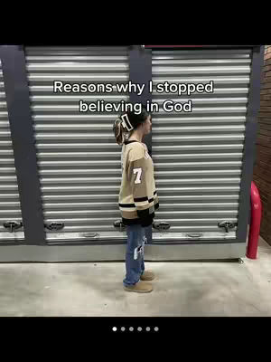 A young woman stands in profile against a backdrop of corrugated metal doors. She is wearing a tan sweater with dark stripes and the number 7, blue jeans, and boots. Text overlayed on the image reads, 'Reasons why I stopped believing in God'.