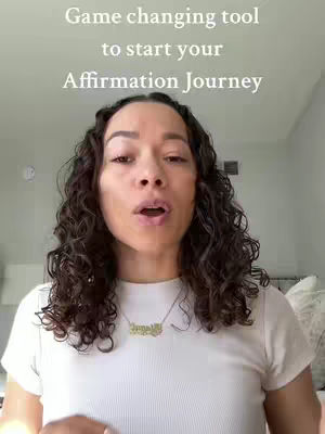 A woman with curly dark hair stands in front of a neutral-colored background. She wears a white t-shirt and a gold necklace. Text overlayed on the video reads, 'Game changing tool to start your Affirmation Journey'.