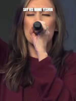 A woman with long light brown hair is holding a microphone and singing with her eyes closed. Text overlayed reads 'SAY HIS NAME YESHUA'. The background is blurred and appears to be a stage with lighting.