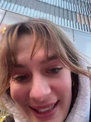 A close-up shot features a young woman with fair skin, light brown hair with bangs, and a white hoodie. Her eyes are visible and are slightly looking up while she is smiling and happy. The background of the image include a metallic paneled building.
