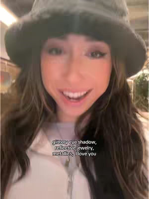 A selfie-style image featuring a young woman with long brown hair wearing a black bucket hat. She is smiling at the camera with lipstick and eye makeup. Text overlay on the image contains describing some makeup details. The background appears to be an indoor setting, possibly a retail store.