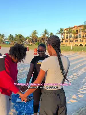 Three women of African descent are holding hands in a circle on a beach. The sky is clear and blue, and the beach has palm trees and buildings in the background. The women are dressed casually, and appears to be a prayer or worship gathering.
