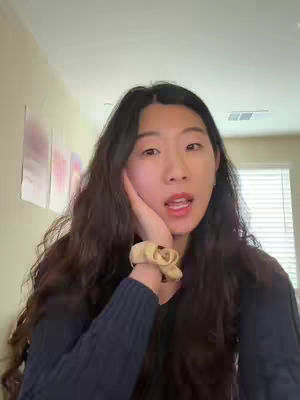 A young woman with long, wavy dark hair is the main focus. She has her hand on her cheek and is looking at the camera with her mouth open. The background is a room with beige walls and some art pieces.