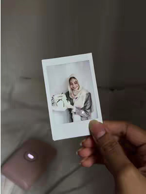 A photo held by hand, featuring a woman with a hijab in a Polaroid frame. The woman holds a bouquet of flowers and smiles at the camera. A pink instax camera is in the background.