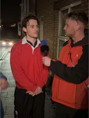 A man in a red rugby top is interviewed by another man outdoors at night. The background shows a dimly lit street.