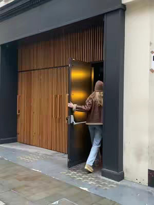 A person with long blonde hair, wearing a brown jacket and hat, opens a door. The door is dark gray, and reveals a brightly lit interior. The exterior of the building features a wooden paneling on the left and a plain, light-colored wall on the right. The ground in front of the door shows a slightly wet or dark surface.