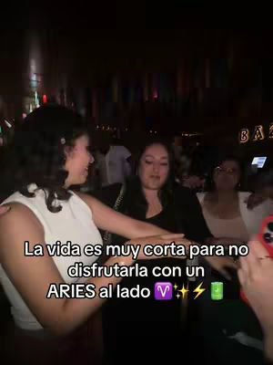 Three women are in a dimly lit environment, likely a bar. One woman, on the left, is wearing a white top and is turned toward the center, smiling. Another woman, in the center, is wearing a black top. The background is blurred, but there are other people present, along with a sign that says "Bar."