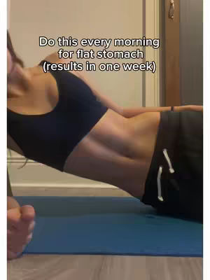 A woman is shown from the abdomen up in a side plank position, emphasizing the core muscles. She is wearing a black sports bra and black athletic shorts. The background shows a plain wall and a sliver of a wooden door frame. Text overlaid on the image proposes a fitness routine for a flat stomach as the exercise continues.