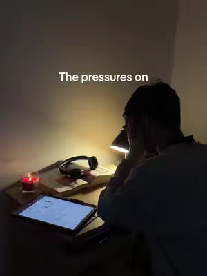 A person sits at a desk leaning forward with their head in hands, appearing stressed. A lamp provides warm light, illuminating the desk and revealing a book, tablet, and headphones.