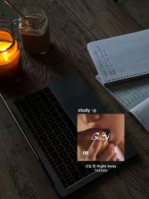 An overhead shot of a wooden desk with a laptop, notebook, and a candle. A music player interface is overlaid on the laptop screen. The scene has warm lighting and is set up to suggest a study environment.